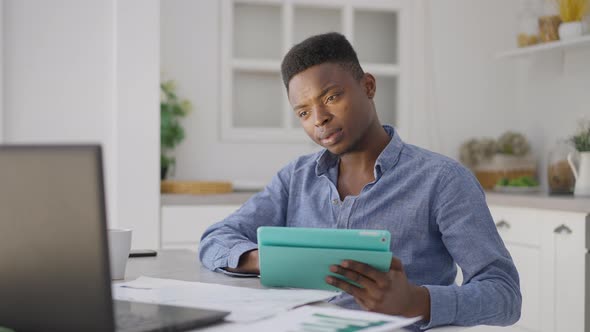 Portrait of Young Handsome Man Scrolling Tablet Screen Checking Laptop Sitting at Kitchen Table