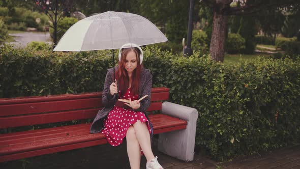 Young Beautiful Woman in Headphones with Transparent Umbrella Reading Book Sitting on Bench in City