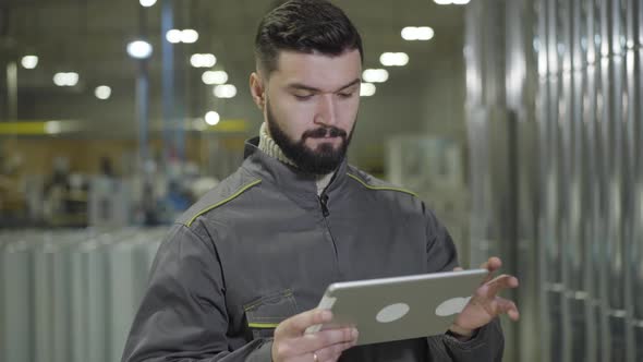 Portrait of Concentrated Bearded Caucasian Man Standing at Production Site and Using Tablet. Modern