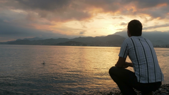 The Man Looks In a Shirt Looking At The Sunrise On The Beach Of The Ocean. The Sun Rises From Behind