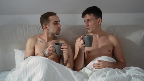 Portrait of Loving Gay Couple Drinking Morning Coffee Sitting in Bed at Home Indoors