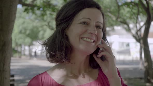 Cheerful Mature Lady Talking on Smartphone in Summer Park