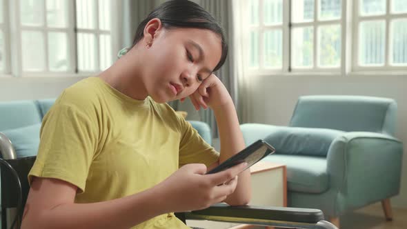Bored Asian Kid Girl Sitting In A Wheelchair While Using Smart Phone At Home alt