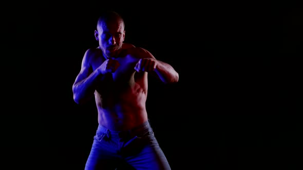 a Muscular Bald Man with a Bare Torso and Jeans Strikes Straight in Front of Him on a Black