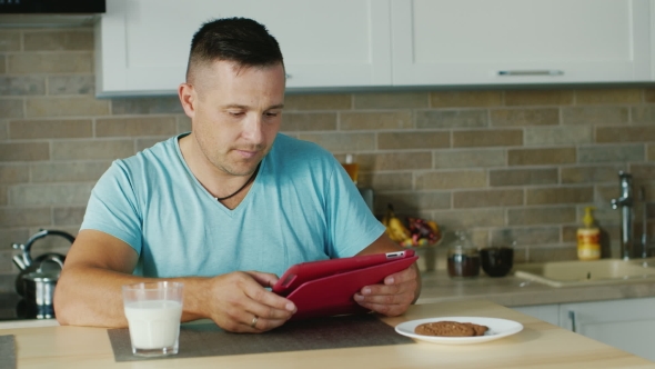 A Young Man Uses Tablet In The Kitchen, Standing Next To a Glass Of Milk. Healthy Lifestyle
