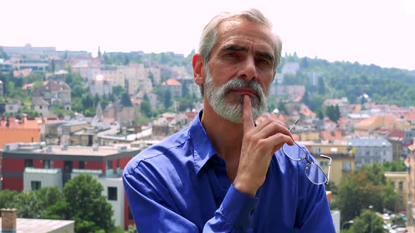 Old Senior Man Walks and Thinks About Something with Glasses in Hand - City in Background