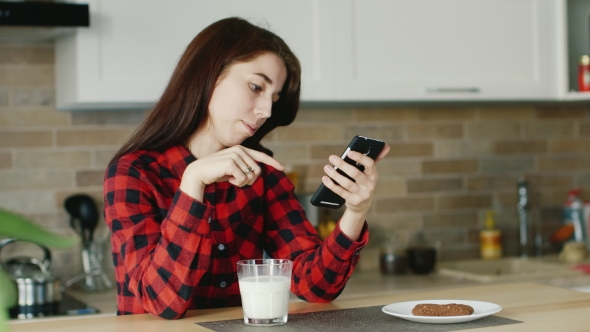 A Young Woman Uses a Smartphone. He Is Sitting In His Kitchen, Standing Next To a Glass Of Milk