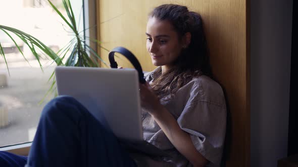 Female Freelancer Sitting on Windowsill in Cafe and Working on Laptop in Headphones