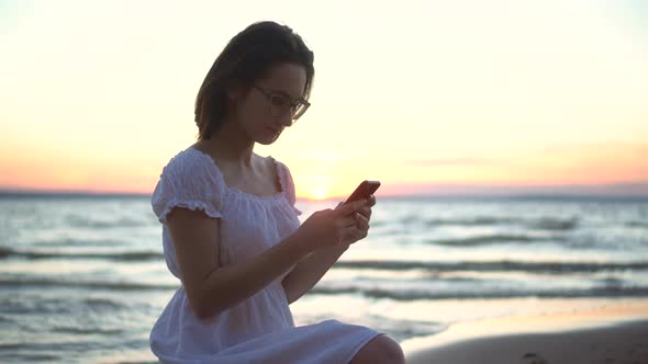 A Young Woman Sits on a Stone on the Beach By the Sea with a Phone in Her Hands. A Girl in a White