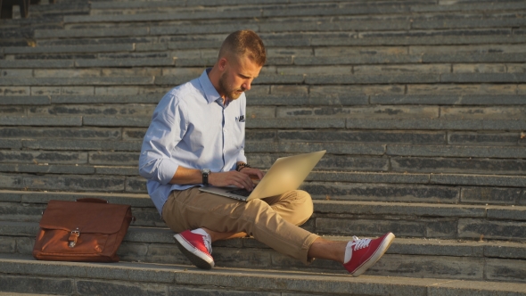 Handsome Student Sitting On Stone Steps And Using a Laptop