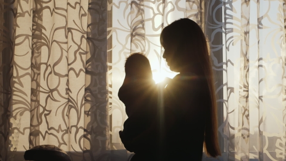 Silhouette Mother With Baby. Standing At The Window At Sunset, The Sun's Rays Shine Beautifully