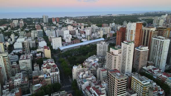 Dolly in flying over Barrancas de Belgrano park and train station surrounded by buildings at sunset,