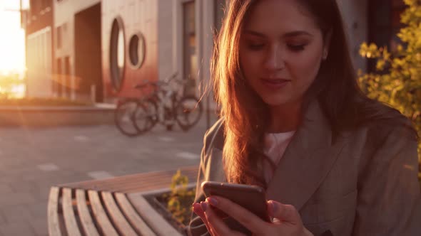 Young attractive business woman sitting outdoor on the bench and using smartphone.