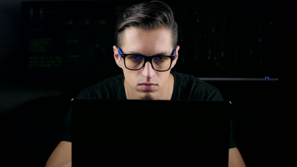 Male Hacker With Glasses Working On a Computer In a Dark Office Room