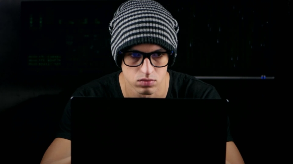Male Hacker With Glasses And Hat Working On a Computer In a Dark Office Room