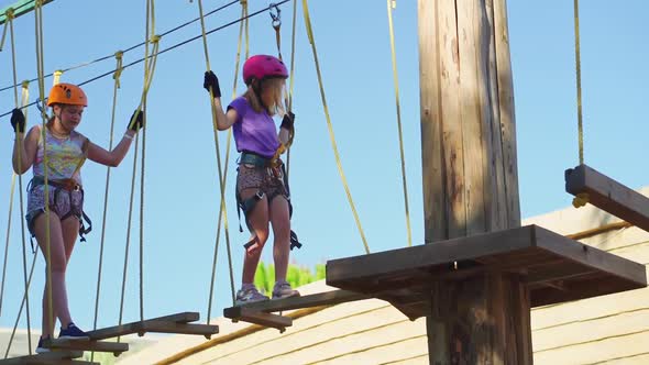 Two Frightened but Brave Girls in Helmet Tshirt and Shorts Climbs in Rope Park