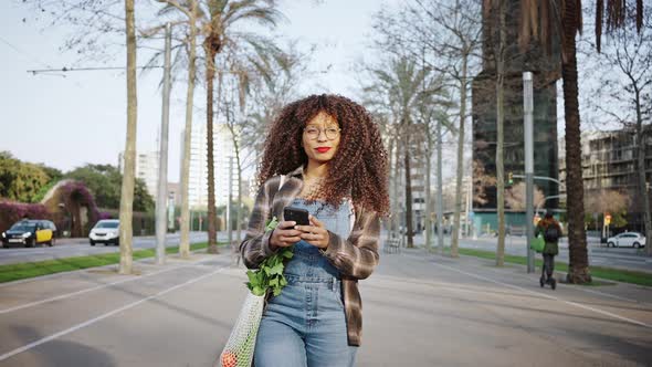 A Girl Wearing Denim Overalls and Glasses Walks Along Streets of Barcelona with a Phone in Her Hands