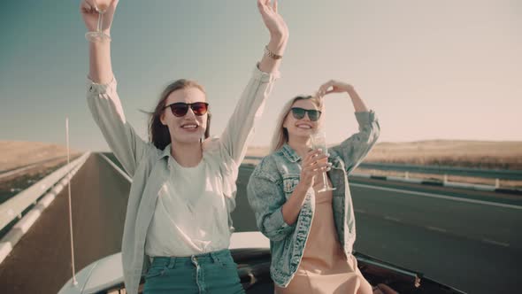 Two happy women with champagne in their glasses ride in convertible on highway.