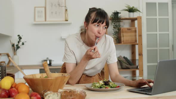 Young Woman Eating Salad and Using Laptop in Kitchen