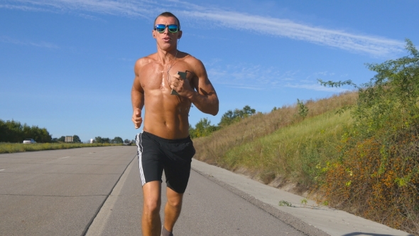 Young Smiling Muscular Man In Sun Glasses Jogging At Highway And Listening Music In Headphones From