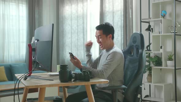Asian Cameraman Looking At Mobile Phone And Being Happy Due To Success While Using Computer at Home