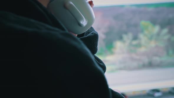 Woman Drinks Coffee From Cup in Black Bathrobe at an Open Window in the Morning