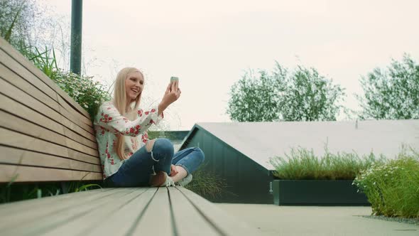 Lovely Young Female Using Smartphone To Make Video Call While Sitting on Bench on Living Roof of