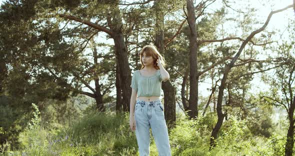 A Cheerful Teenage Girl with Curly Hair Stands in a Clearing Between Green Trees in Summer Forest