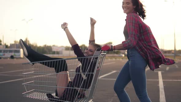 Side View of a Happy Young Woman Pushing a Grocery Cart with Her Girlfriend Inside in the Parking By