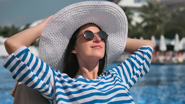 Relaxed Smiling Woman in Sunglasses and White Hat Sunbathing Near Swimming Pool Medium Closeup