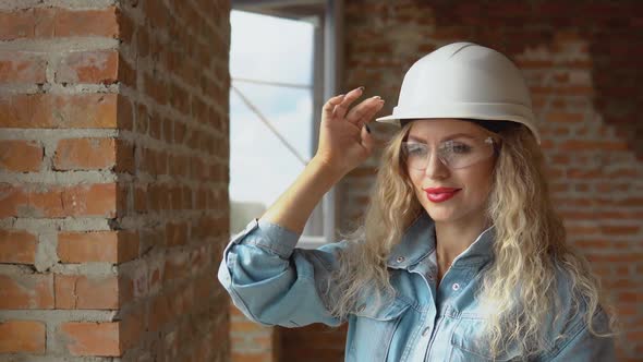 A Young Woman in a White Work Helmet and Denim Clothes and Goggles Stands at the Construction Site