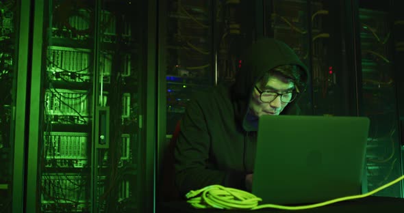 Asian male hacker in hoodie using laptop by computer servers