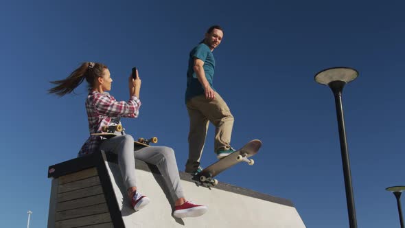 Happy caucasian woman taking photo of her male friend skateboarding