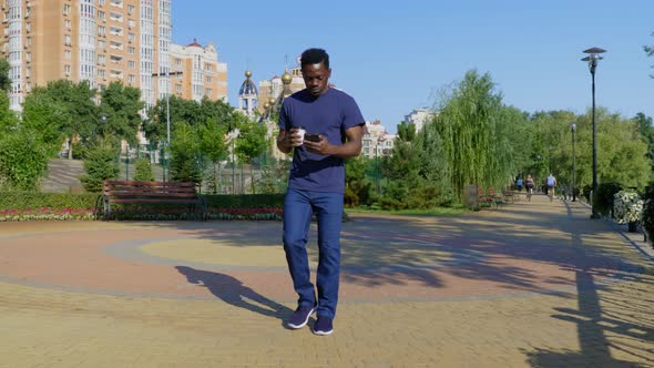 Smiling Afroamerican Man Walks Street Uses Mobile Phone Holding Cup with Coffee