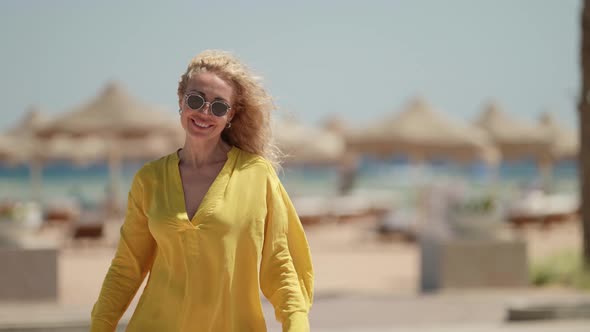 a Blonde in Sunglasses and a Yellow Fluttering Dress Smiles and Ruffles Hair Against the Background