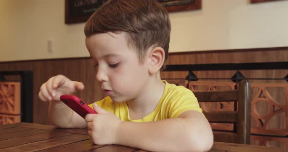 Portrait of a Little Boy Child with a Beautiful Face Looking at the Phone While Sitting at the Table