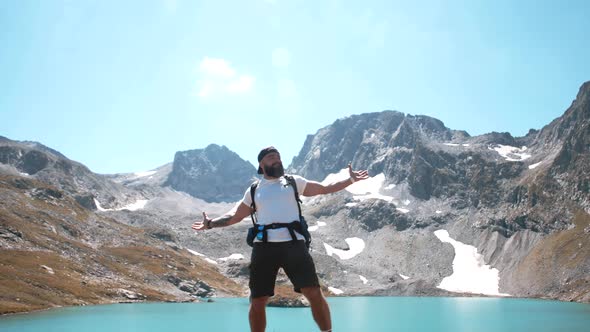 A young hiker with a beard stands on the top of a mountain near a mountain lake