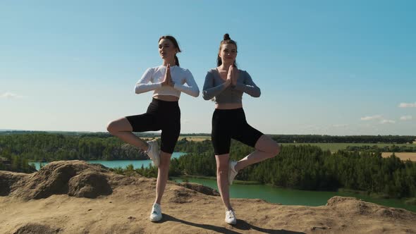 Two Athletic Women in Tops and Sporty Shorts Stand in Yoga Pose on the Edge of Mountain Against the