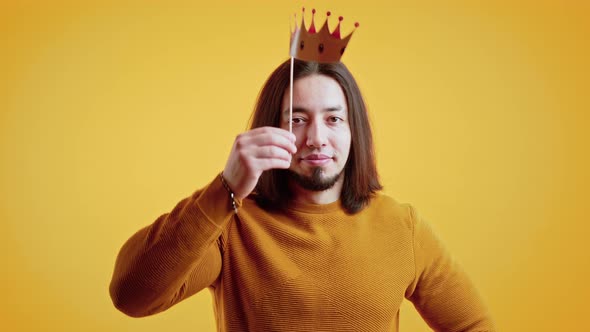 Caucasian Man with Long Brown Hair Wearing Orange Sweater Holding Paper Crown Above Head and