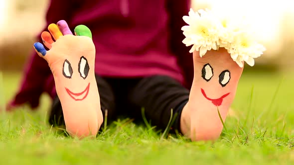 Little Child Girl Sits on Grass with Painted Feet with Funny Comic Faces