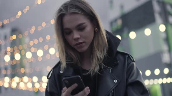 Young Girl Uses a Smartphone Sitting Outside in a Modern Residential Area Evening in the City Blurry