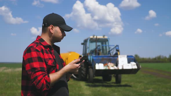 Young Attractive Farmer with Phone Standing in Field, Tractor Working in Green Field in Background