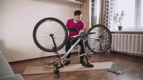 Concentrated Man Sits with Tablet in Room Near Inverted Bicycle