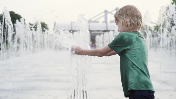 Little boy plays in the square between the water jets in the city fountain at sunny summer day