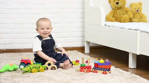 a Baby in a Denim Jumpsuit Sitting Barefoot on the Carpet Smiles and Plays with Toys