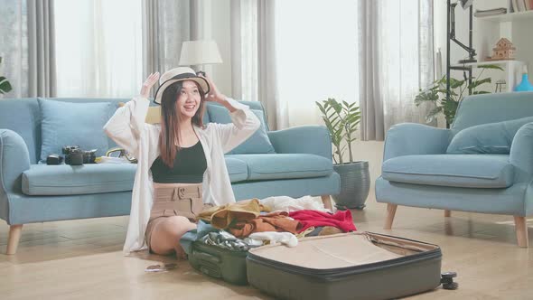 Asian Young Woman Try Hat And Packing Clothes In Suitcase At Home, Preparing For Vacation