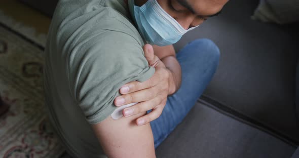 Young man in protective face mask sitting on sofa after vaccination during coronavirus outbreak