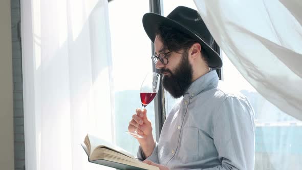 Pleased Bearded Man Drinking Red Wine While Reading a Book at Home