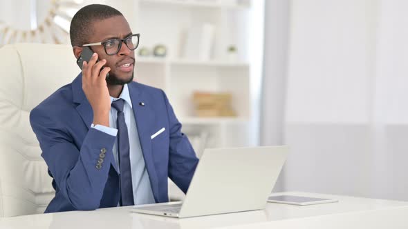 African Businessman with Laptop Talking on Smartphone in Office 