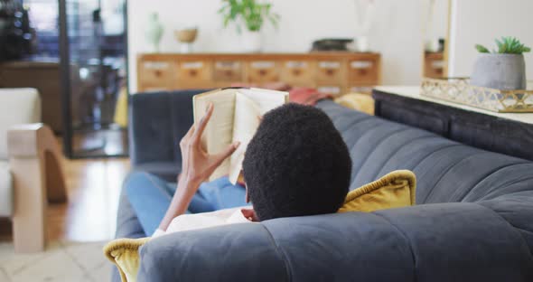 Happy african american woman lying on sofa in living room, reading book